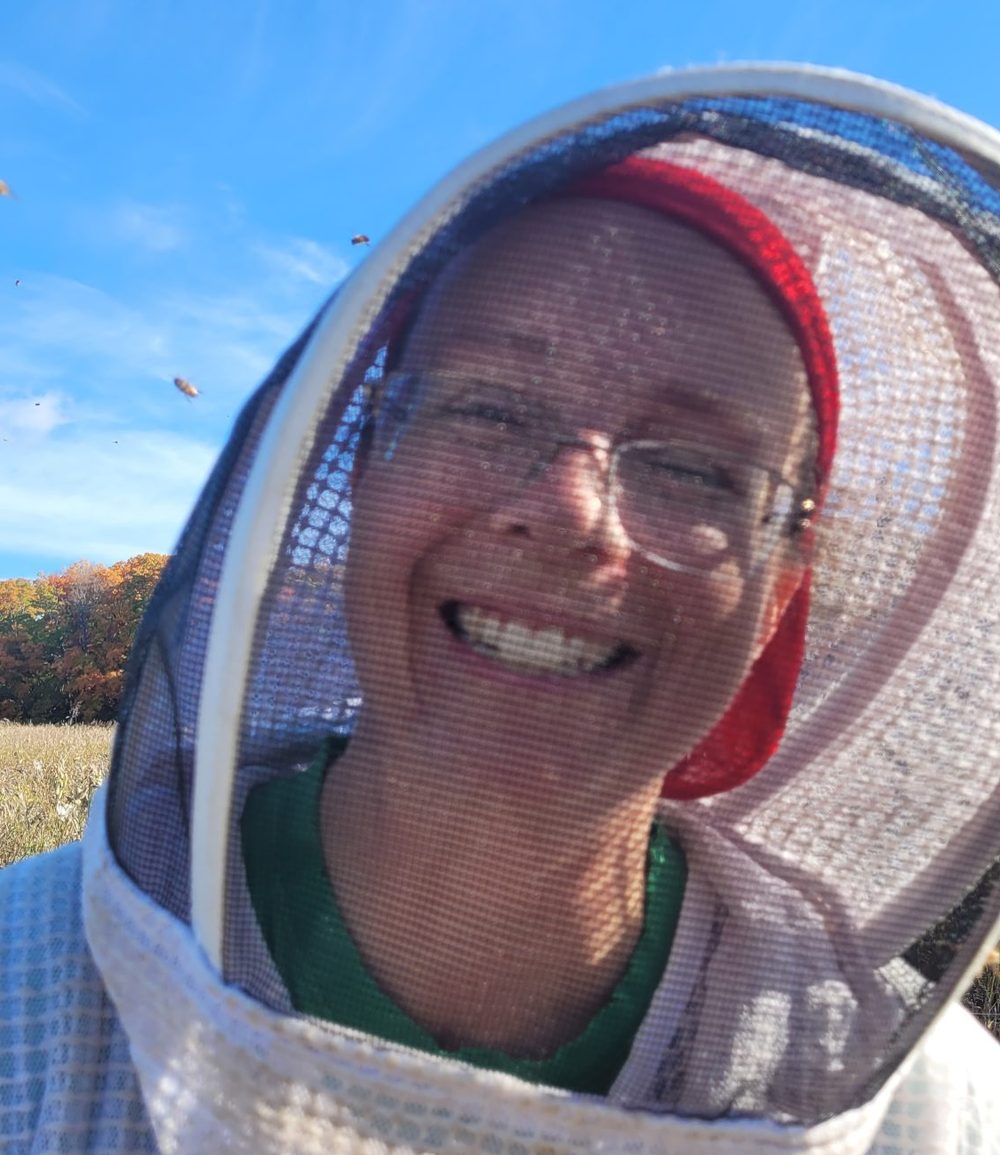 Lisa St Denis smiling through her bee veil on a sunny autumn day, with bees in the air behind her.